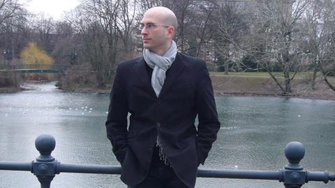 Der Komponist Peter Gahn, seit 2015 Professor für Komposition/Neue Medien/Sound Studies an der Hochschule für Musik Nürnberg, steht vor einem Geländer an einem kleinen Fluss