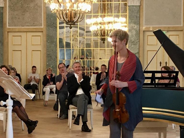 Eine Frau mit kurzen, blonden Haaren hält eine Geige in der linken Hand und steht in einem Schlosssaal vor einem Cembalo. Um sie herum verteilt sitzen Menschen mit viel Abstand und applaudieren.