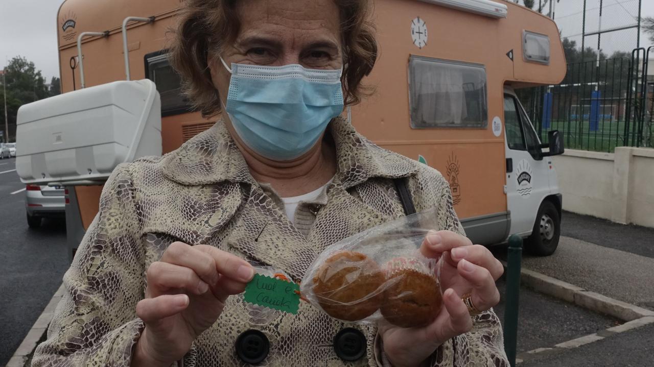 Eine Frau mit blauer Mund-Nasen-Maske hält ein Tütchen mit zwei kleinen Kuchen in der Hand, daran hängt ein grüner handgeschriebener Zettel mit der Aufschrift "Obrigada" (Portugiesisch: "danke").
