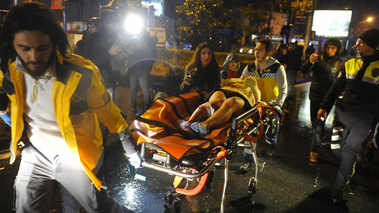 Zwei Sanitäter rollen die am Bein verletzte Frau auf einer Trage über eine Straße. Dahinter laufen weitere Menschen.