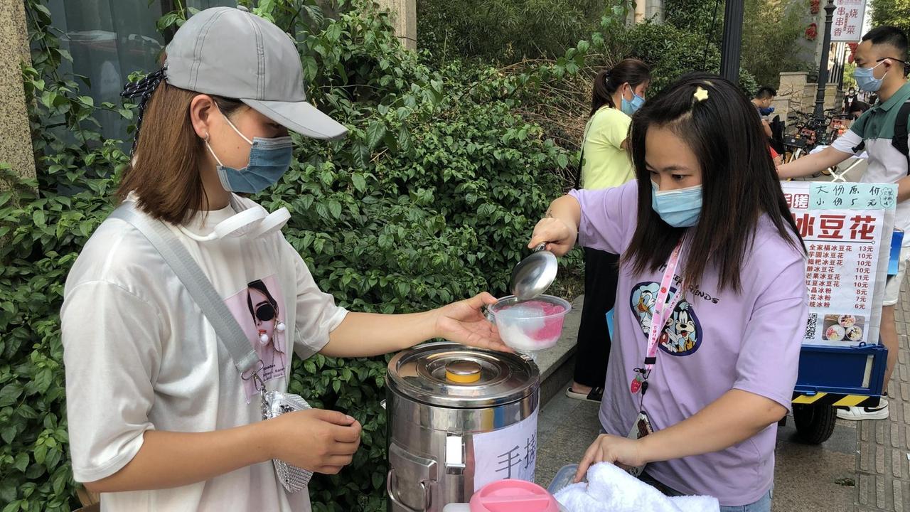 Eine junge Frau hält an einem Straßenstand ein Gefäß aus Plastik, in das eine andere Essen füllt.