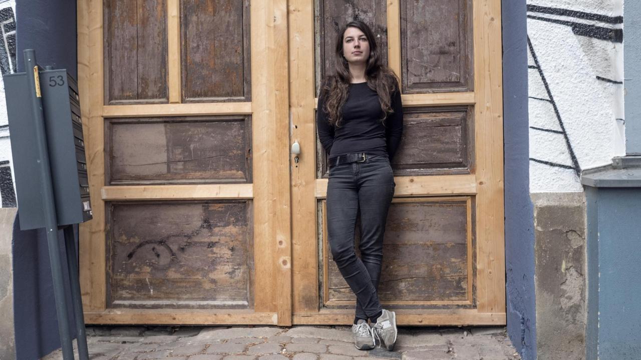 Luise Grudzinski. Eine junge Frau steht an einer Holztür eines Gebäudes.