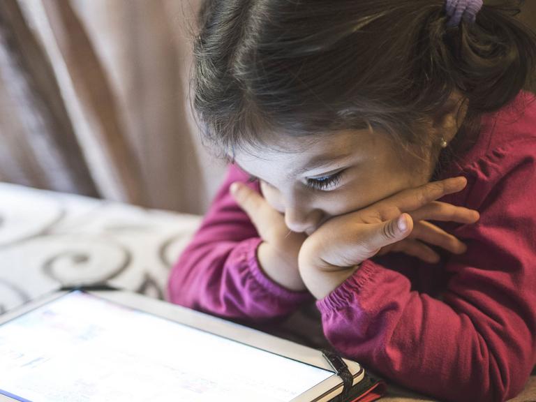 Ein kleines Mädchen stützt sich mit den Armen auf ein Bett und schaut auf ein Tablet.