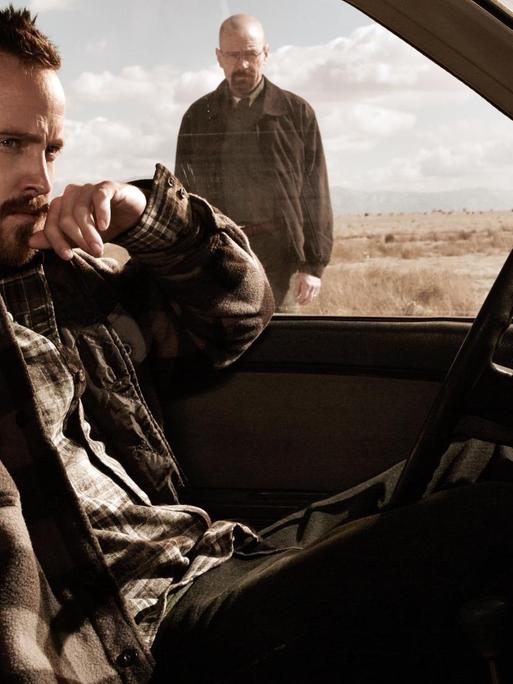 Szenenbild aus "Breaking Bad": Aaron Paul als Jessie Pinkman sitzt im Auto, im Hintergrund sieht man Bryan Cranston als Walter "Walt" Hartwell White stehen.
