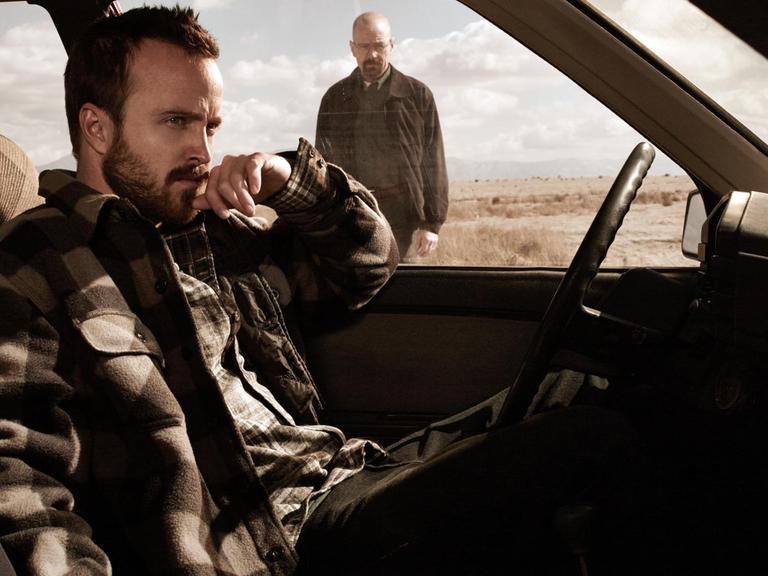 Szenenbild aus "Breaking Bad": Aaron Paul als Jessie Pinkman sitzt im Auto, im Hintergrund sieht man Bryan Cranston als Walter "Walt" Hartwell White stehen.