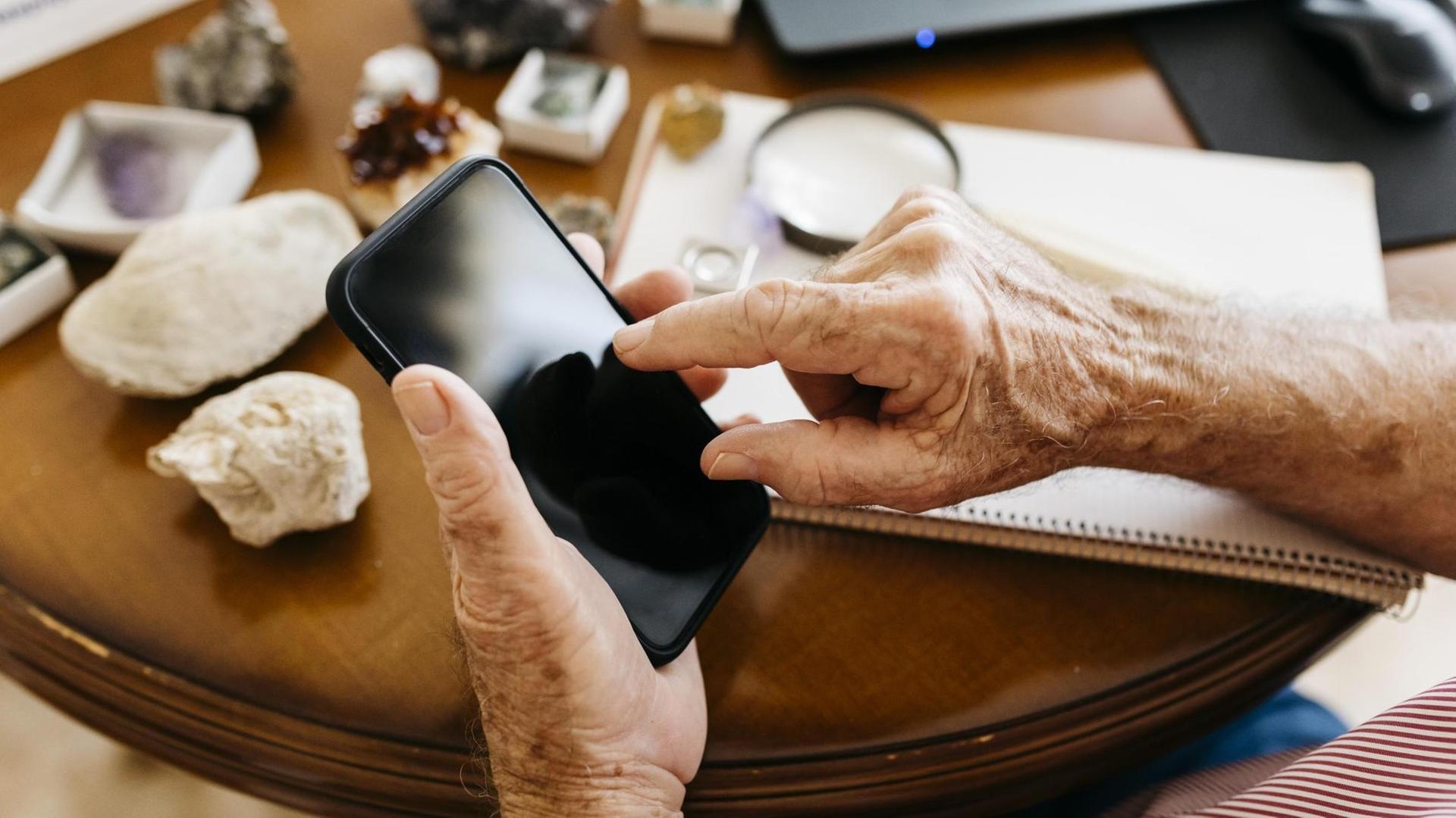 Hände eines älteren Mann, der ein Mobiltelefon in der Hand hält.
