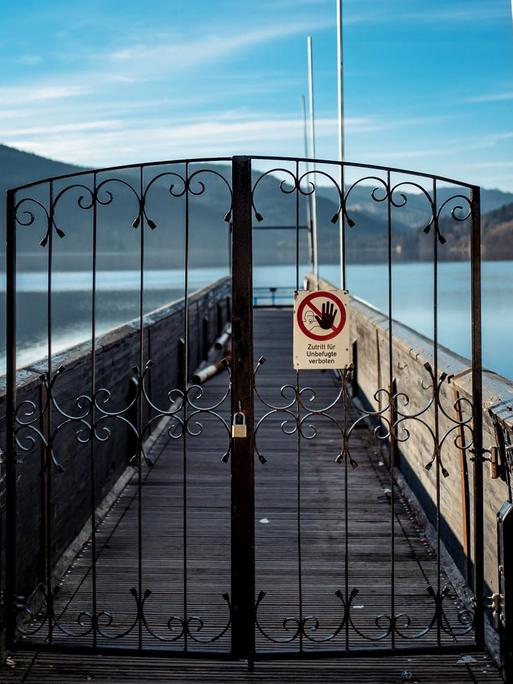Blick auf einen Steg, der auf einen sonnenbeschienenen See führt. Davor ein verschlossenes schmiedeeisernes Tor mit einem Schild, auf dem steht "Zutritt für Unbefugte verboten".