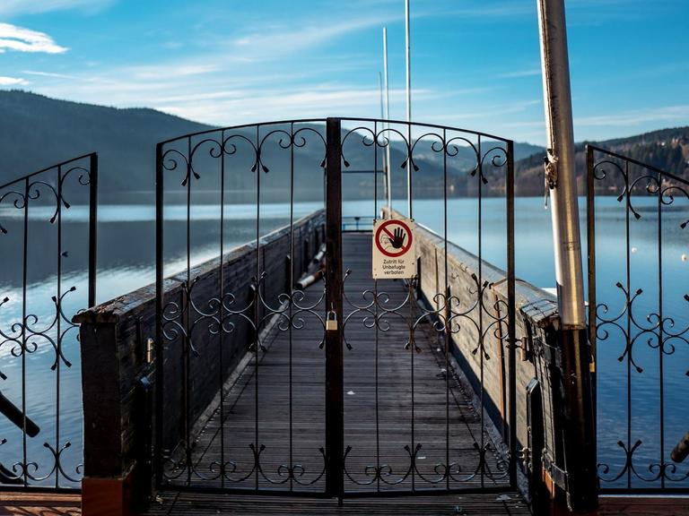 Blick auf einen Steg, der auf einen sonnenbeschienenen See führt. Davor ein verschlossenes schmiedeeisernes Tor mit einem Schild, auf dem steht "Zutritt für Unbefugte verboten".