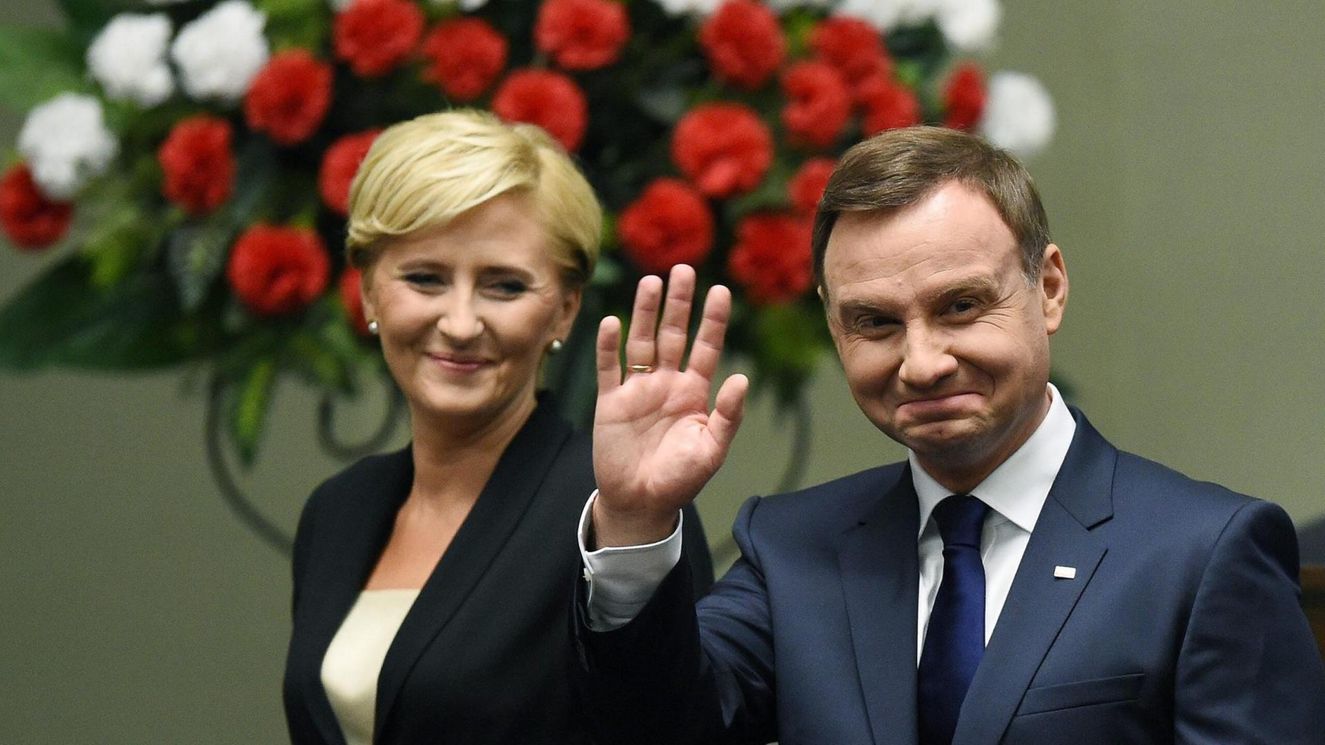 Polens Präsident Andrzej Duda winkt bei seinem Amtsantritt in Warschau in die Kameras. Der Präsident hat kurze braune Haare und trägt einen blauen Anzug, ein weißes Hemd und eine dunkle Krawatte. Links neben ihm steht seine Frau Agata Kornhauser-Duda, sie trägt eine blonde Kurzhaarfrisur, einen schwarzen Blazer, darunter ein weißes Oberteil. Im Hintergrund sieht man rot und weiß blühende Blumen.