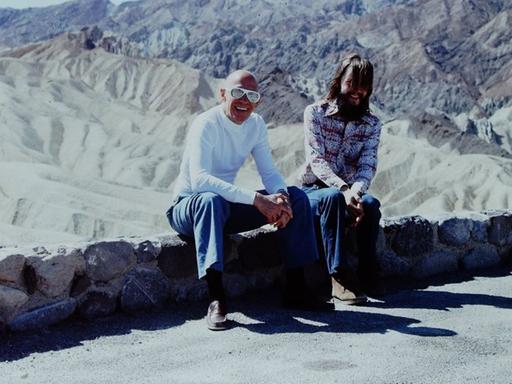 Am Rande einer Strasse auf einem kleinen Mäuerchen sitzen Foucault und Stoneman. Im Hintergrund die Berge vom Death Valley.