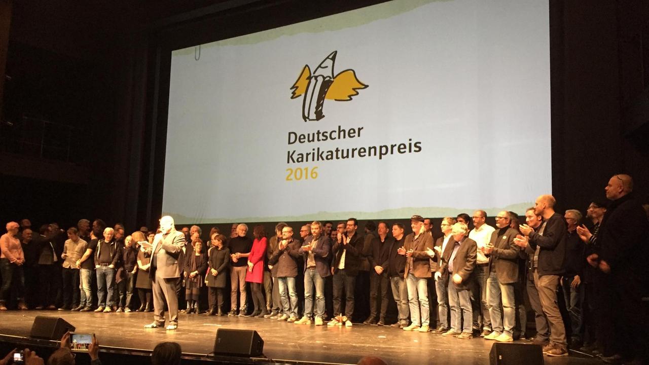 Die Teilnehmer des deutschen Karikaturistenpreises 2016 auf der Bühne. Auf der Leinwand zu sehen: Der geflügelte Bleistift.