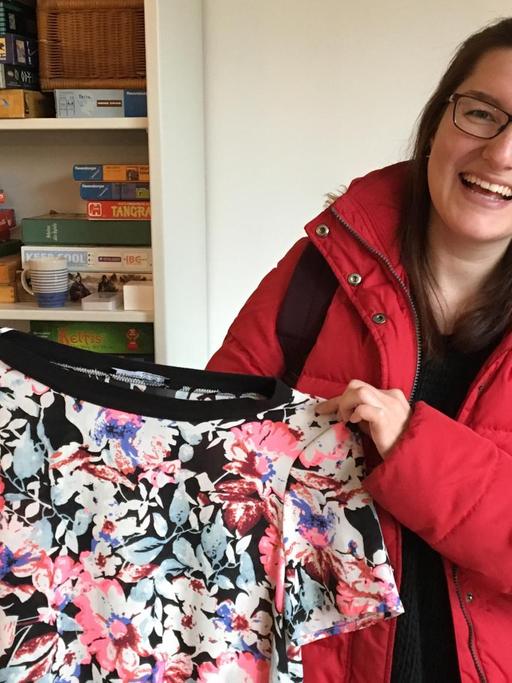 Eine junge Frau mit langen dunklen Haaren, Brille und roter Steppjacke steht vvor einem Regal mit Büchern und Gesellschaftsspielen und hält lachend ein geblümtes T-Shirt in die Kamera.