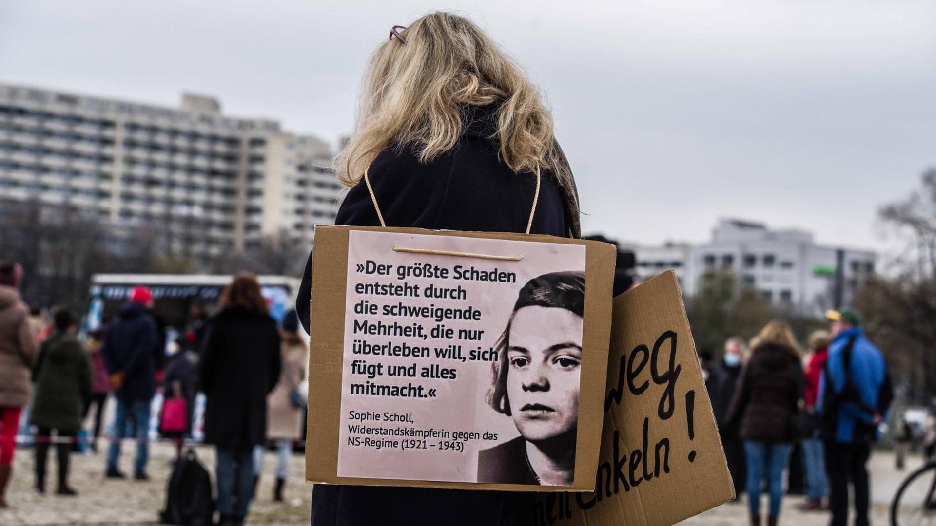 Rückansicht einer Frau auf einer Demo. Sie trägt um den Hals ein Schild mit dem Foto von Sophie Scholl, auf dem das Scholl zugeschrieben Zitat steht: "Der größte Schaden entsteht durch die schweigende Mehrheit, die nur überleben will, sich fügt und alles mitmacht."