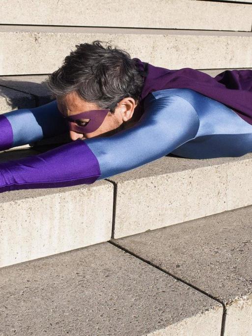 Ein Mann als Superheld verkleidet, mit violetter Augenbinde und Handschuhen, liegt auf einer Treppe auf dem Bauch, seine Arme nach vorn ausgestreckt.