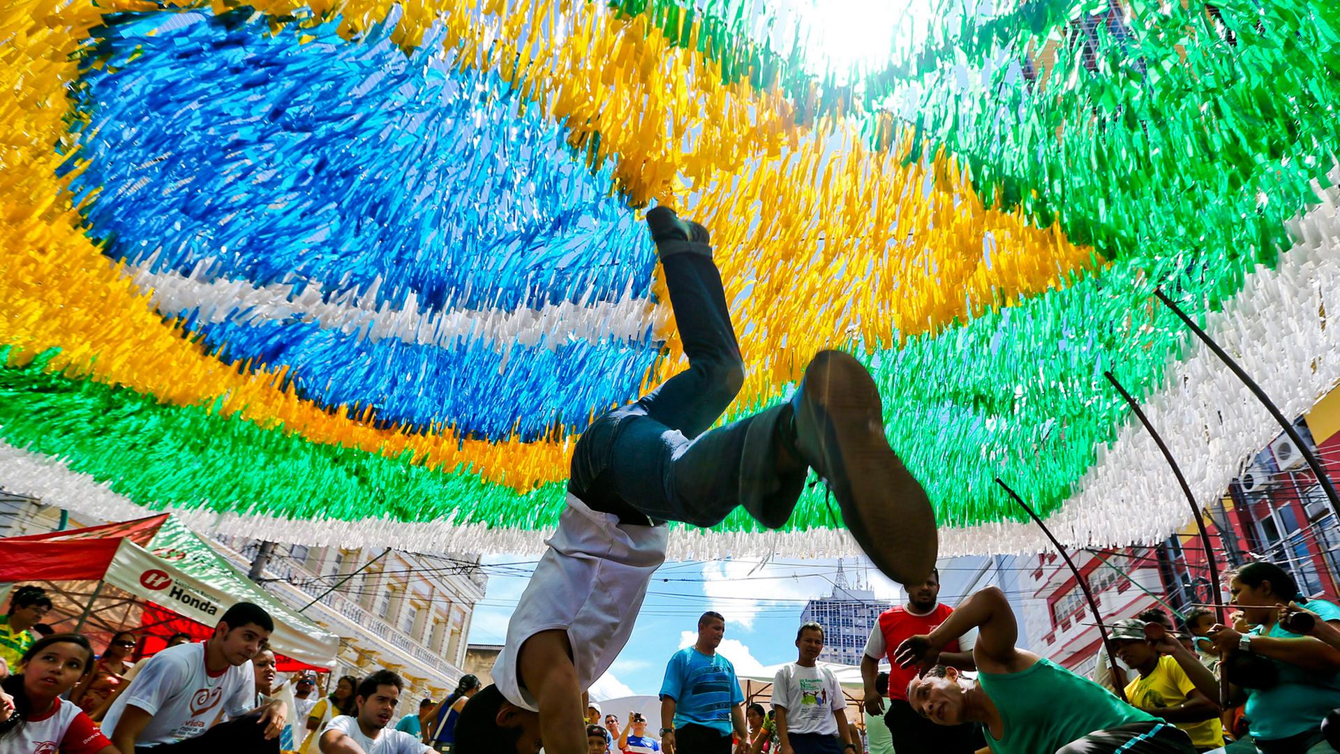 Unter einer brasilianischen Flagge tanzen zwei junge Männer Capoeira: Der eine steht gerade auf seinen Händen, der andere dreht sich am Boden um sich selbst.