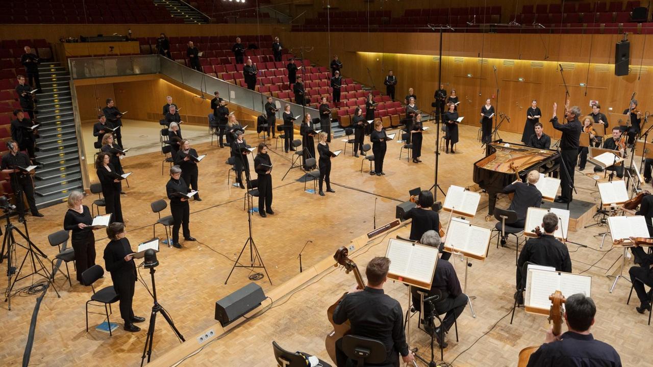 In einem großen Saal sitzt das Orchester hinter dem Dirigenten, der sich gerade den Sängern zuwendet, die im Zuschauerraum verteilt stehen. Zwischendrin stehen Kameras.