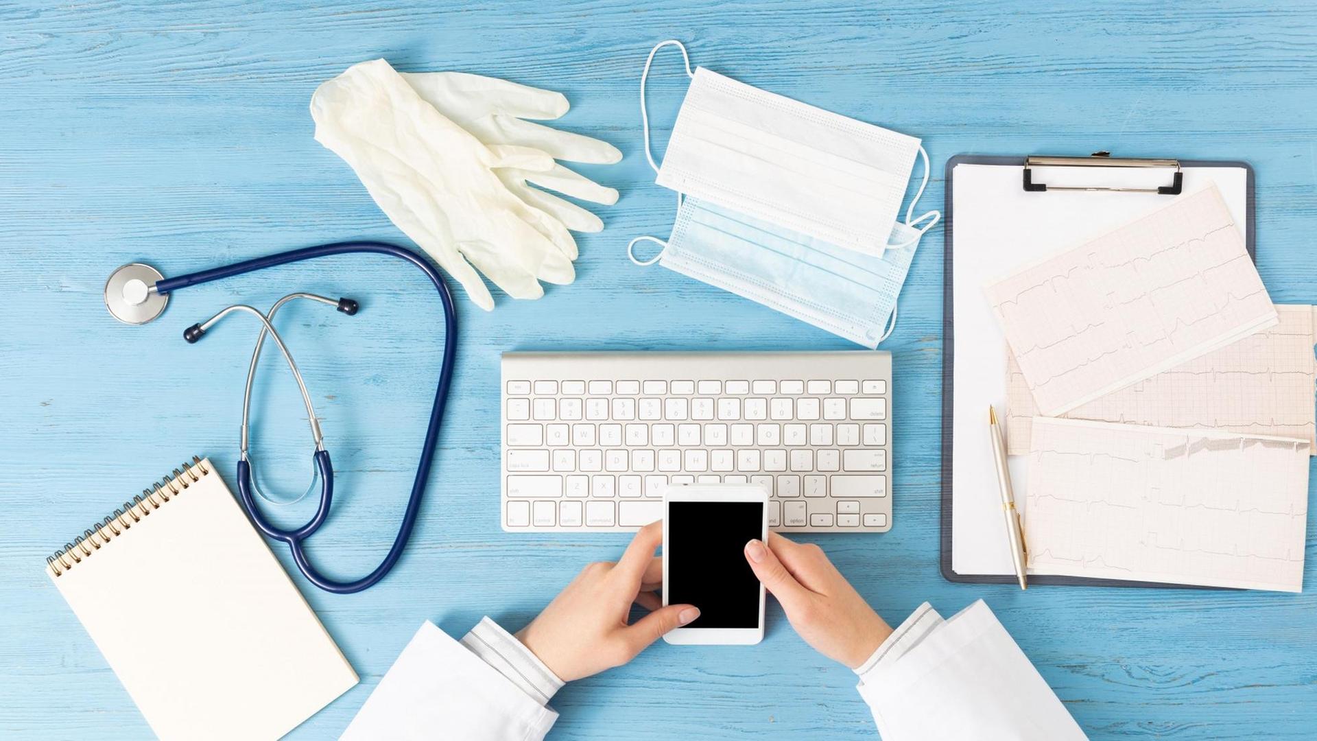 Der Schreibtisch einer Arztpraxis mit Handschuhe, Smartphone, Stereoskope, Maske und Papieren.