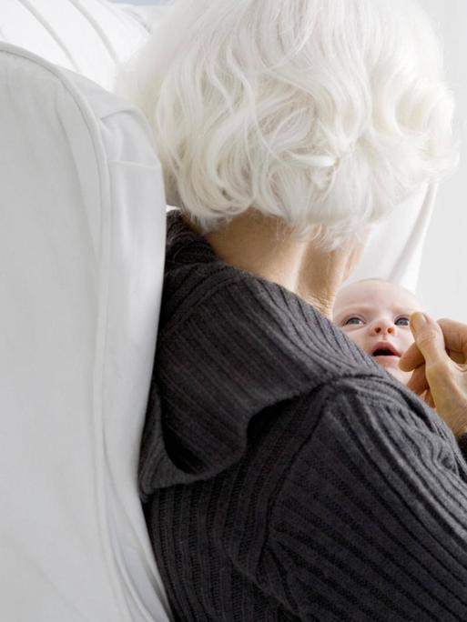 Eine weißhaarige Frau sitzt mit dem Rücken um Betrachter und hält ein Baby im Arm.
