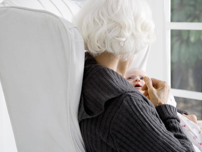 Eine weißhaarige Frau sitzt mit dem Rücken um Betrachter und hält ein Baby im Arm.