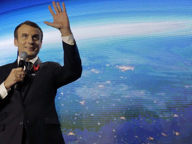 Macron steht lächelnd vor einer großen Videoleinwand mit der Erdkugel, lächelt, spricht und gestikuliert mit der Hand.