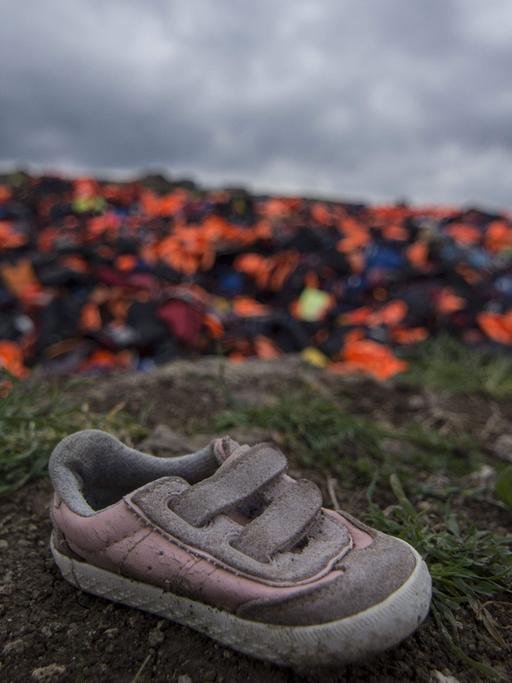 Tausende von Schwimmwesten, die von Flüchtlingen genutzt wurden, stapeln sich auf einer Wiese auf Lesbos. Im Vordergrund ein schmutzig-rosa Kinderschuh.