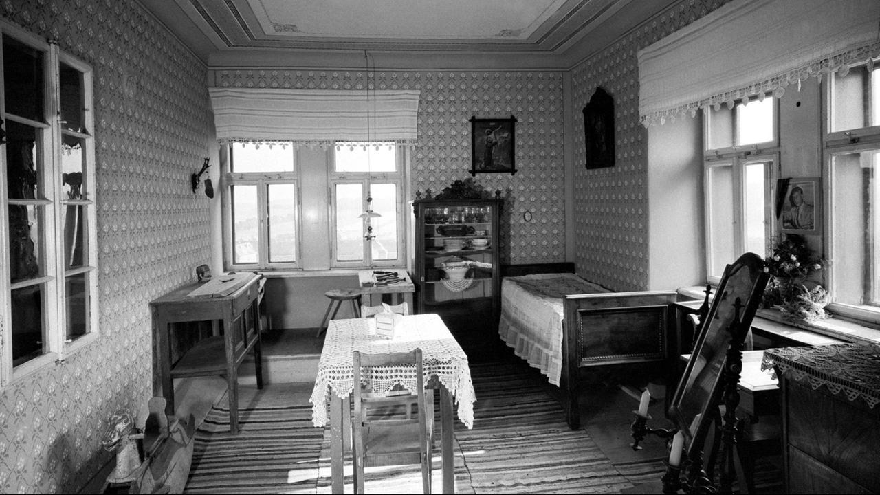 Auf einem schwarz-weiß Foto ist eine kleine Stube zu sehen, in der Bett und Tisch neben einem Küchenregal stehen.