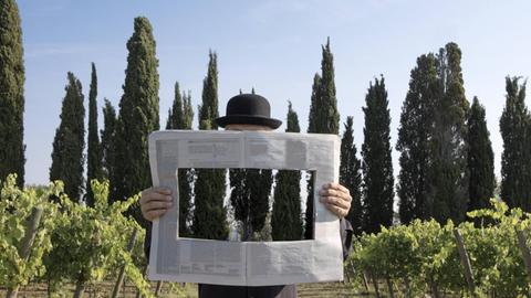 Ein Mann hält eine Zeitung in der Hand, die in der Mitte ein Loch hat, durch das man die toskanische Landschaft im Hintergrund sieht