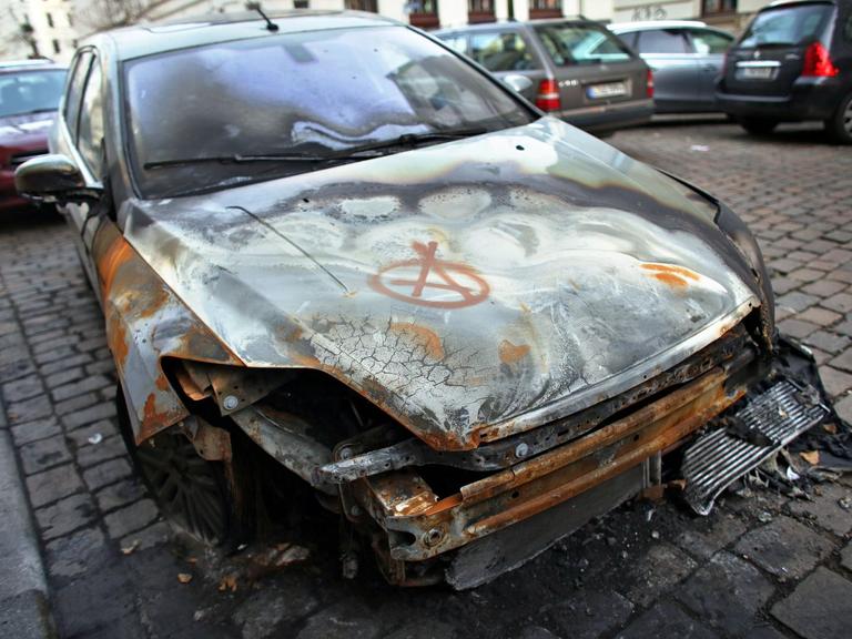 Das Wrack eines von einem Feuer zerstörten Autos steht am 03.12.2013 in Leipzig (Sachsen) in einer Nebenstraße. Darauf ist das Anarchie-Zeichen gesprüht.