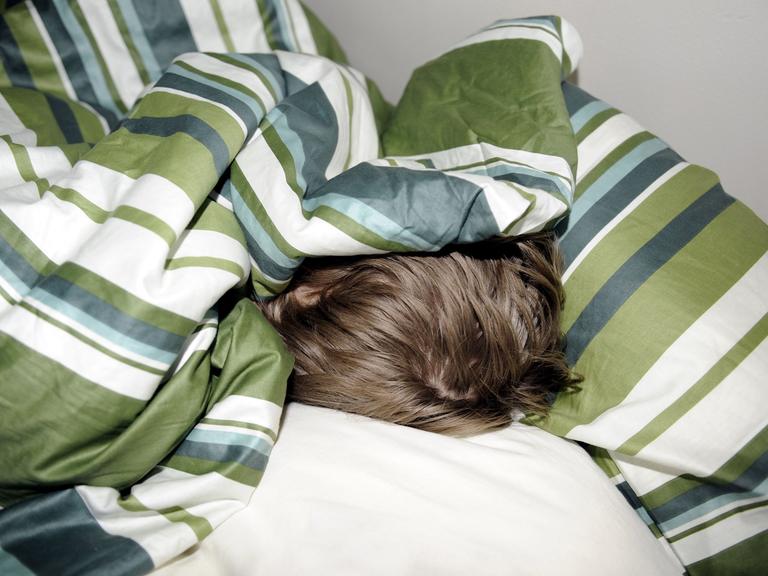 Ein Junge schläft versteckt unter einer gestreiften Bettdecke, nur sein Haarschopf ist zu sehen.