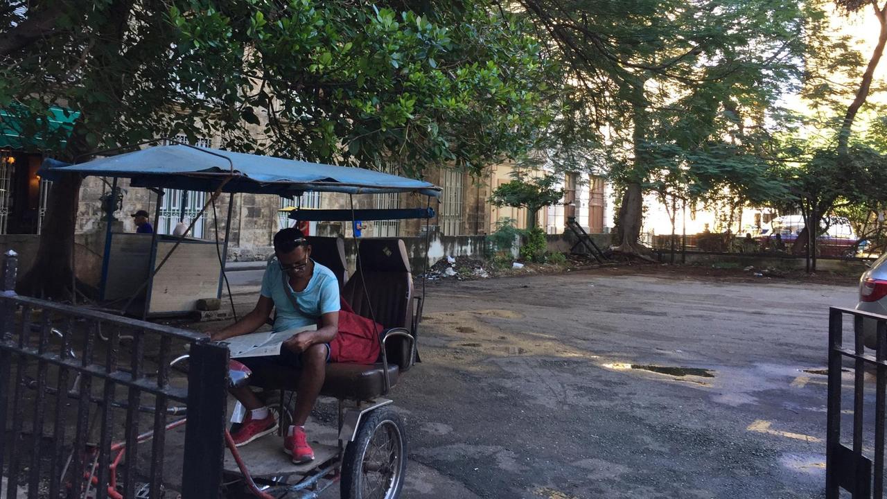 Auf einem schattigen Platz sitzt ein Zeitung lesender Kubaner auf seiner Rikscha und waret auf Kunden.leist ein
