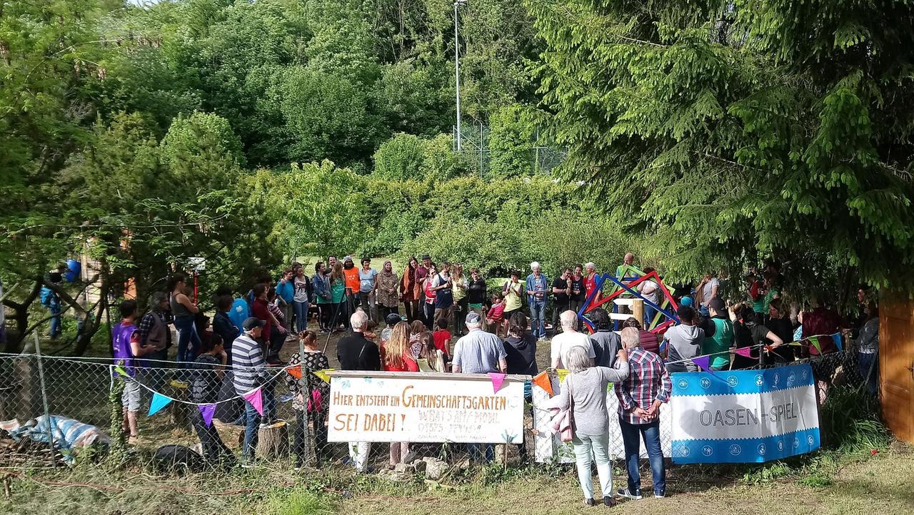 Eine Menschengruppe hat sich in einem Garten versammelt, an dessen Zaun ein Schild hängt mit der Aufschrift: "Hier entsteht ein Gemeinschaftsgarten. Sei dabei!"