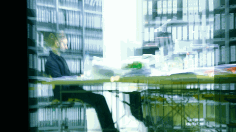Ein Mann sitzt in einem Büro an einem vollen Schreibtisch und arbeitet.
