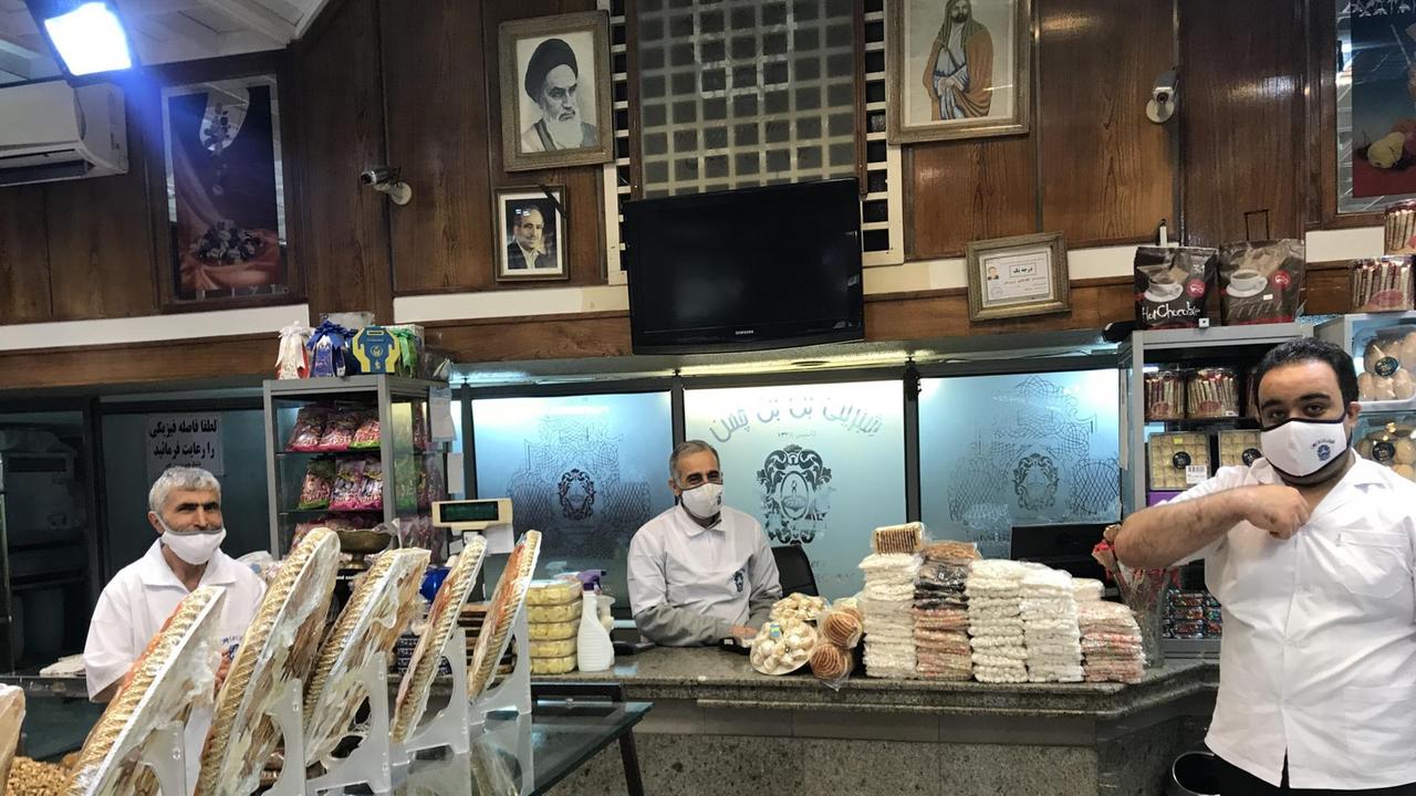 Drei Männer stehen in einer Bäckerein hinter aufgetürmten Backwaren und tragen weiße Hemden und Masken. An der Wand ein Bildnis des Revolutionsführers Ayatollah Khamenei.