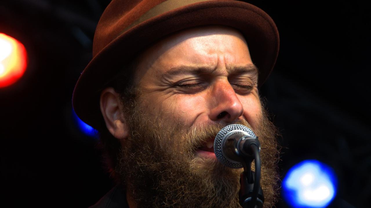 Ein Mann mit Hut und Vollbart steht mit geschlossenen Augen am Mikrofon und singt.