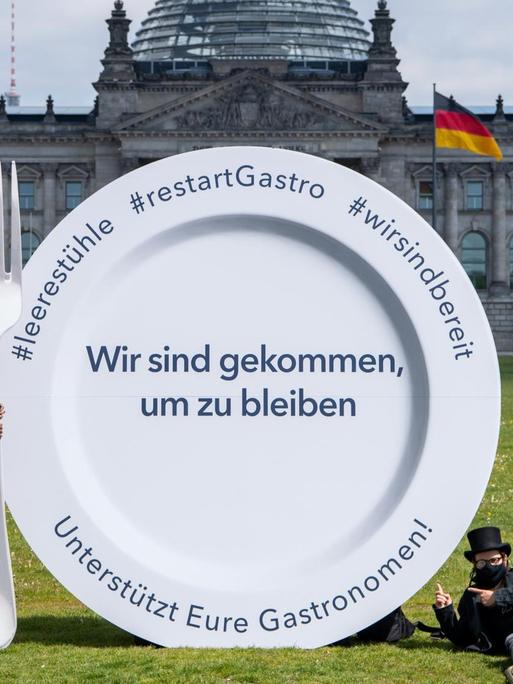 Vertreter des Gastronomie-Gewerbes stehen neben einem großen Teller mit der Aufschrift "Wir sind gekommen, um zu bleiben. Unterstützt Eure Gastronomen!" vor dem Deutschen Bundestag.