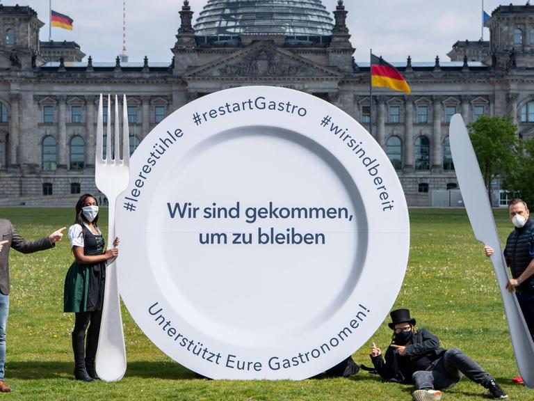 Vertreter des Gastronomie-Gewerbes stehen neben einem großen Teller mit der Aufschrift "Wir sind gekommen, um zu bleiben. Unterstützt Eure Gastronomen!" vor dem Deutschen Bundestag.