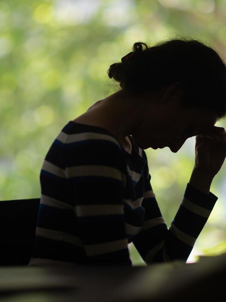 Die Silhouette einer Frau vor einem Fenster. Sie hat den Kopf in die Hand gestützt, ihre Haltung wirkt eingefallen.