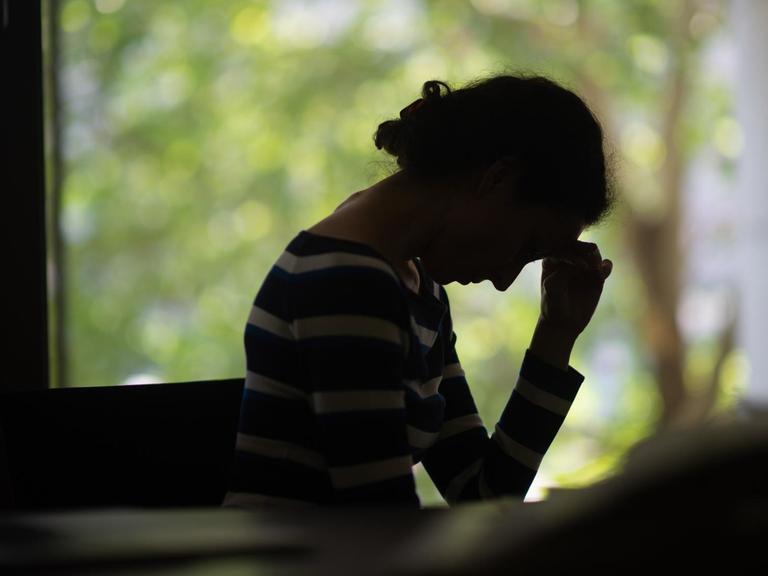 Die Silhouette einer Frau vor einem Fenster. Sie hat den Kopf in die Hand gestützt, ihre Haltung wirkt eingefallen.