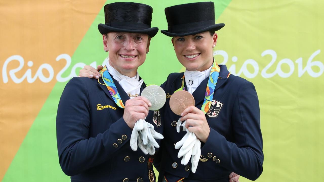 Die beiden Reiterinnen in voller Montur zeigen vor einem bunten Backdrop mit der Aufschrift "Rio 2016" lächelnd ihre Medaillen.