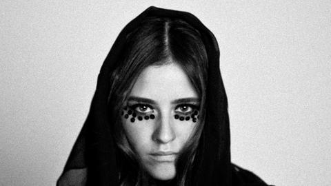 Die brasilianisch-deutsche Musikerin Dillon, mit schwarzer Kapuze und schwarzem Make-Up unter den Augen, schwarz-weiß-Aufnahme, in die Kamera blickend