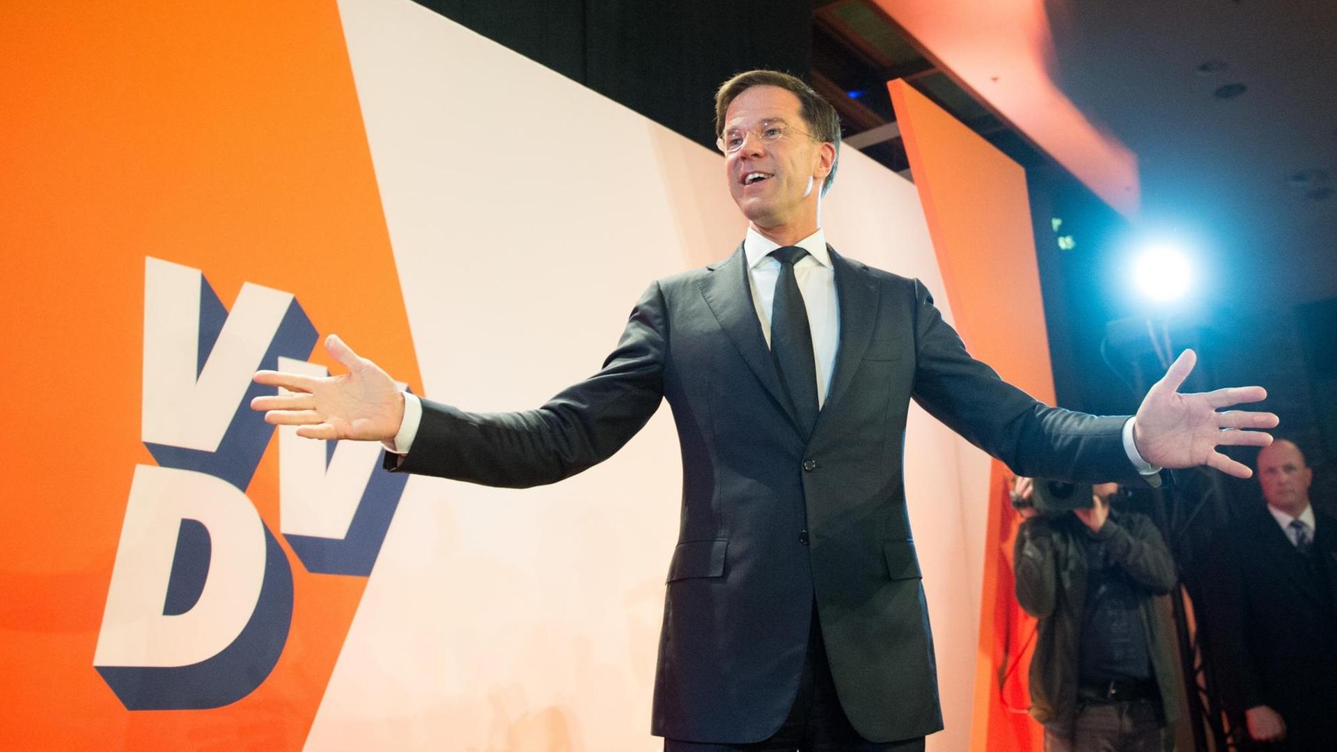 Mark Rutte steht vor einer Wand mit dem Parteilogo der VVD und breitet beide Arme aus.