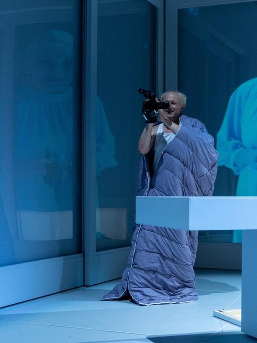 Auf einer hellblau ausgeleuchteten Theaterbühne filmt ein Mann eine Frau aus gewisser Entfernung. Ihr Bild wird im Hintergrund an die Wand projiziert.