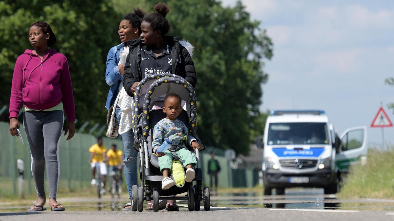 Drei Frauen und ein Kinderwagen auf der Straße vor einem bayerischen Transitzentrum für Flüchtlinge, im Hintergrund Radfahrer, weitere Menschen und ein Mannschaftswagen der Polizei