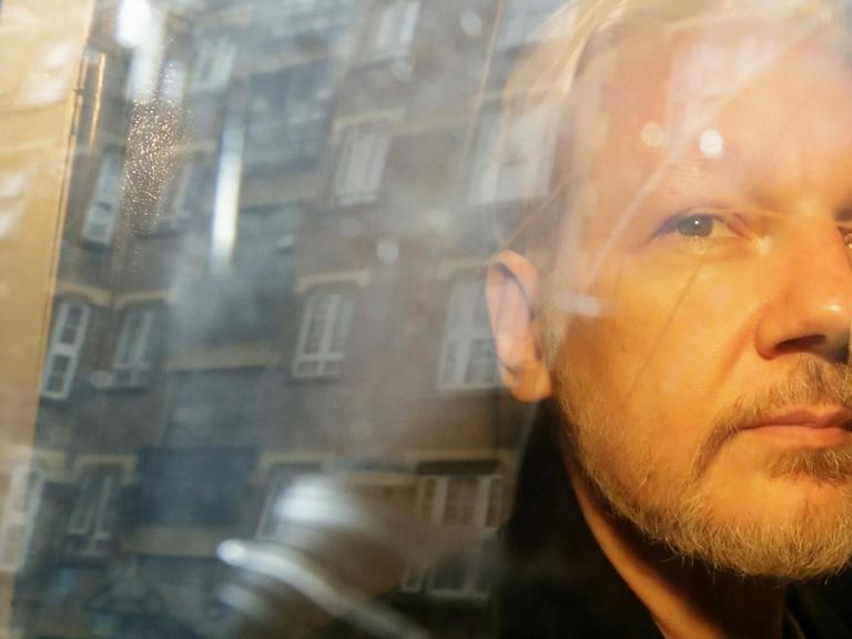 Julian Assange blickt mit gestutztem Bart und streng frisierten Haaren aus einem Busfenster, in dem Gebäude reflektieren.