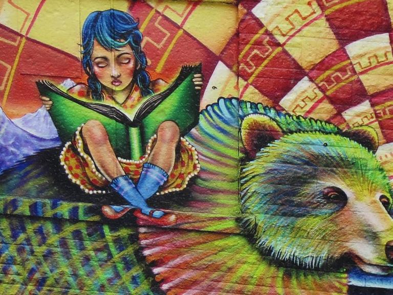 Eine Wandmalerei am Kensington Market in Toronto zeigt ein junges Mädchen, das ein Buch in der Hand hält und liest, während es auf dem Rücken eines Bären sitzt. Der Bär ist sehr bunt, wie auch der ganze Hintergrund.