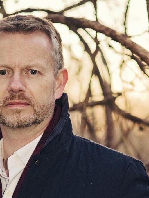 Adam Brookes posiert vor winterlich kahlen Bäumen.