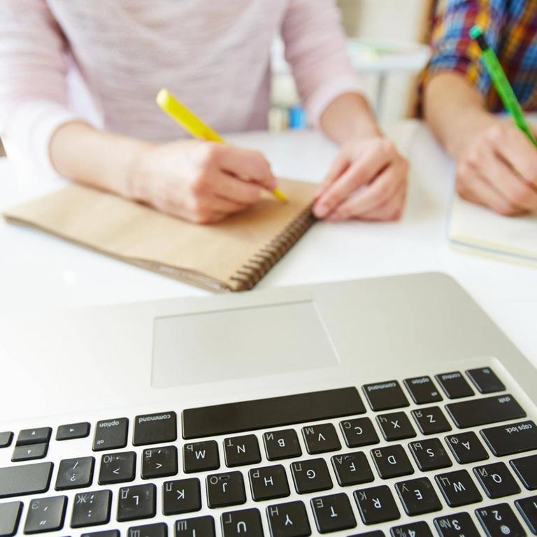 Zwei Schüler schreiben auf ihrem Notitzblock. Vor ihnen steht ein aufgeklappter Laptop.
