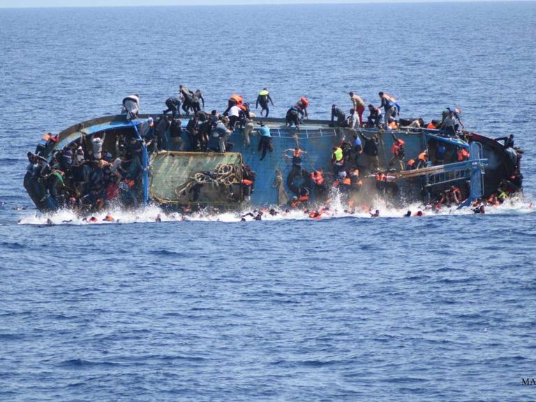 Ein Boot mit Flüchtlingen steht auf der Seite im Wasser, die Menschen fallen und springen zum Teil vom Boot ins Wasser.