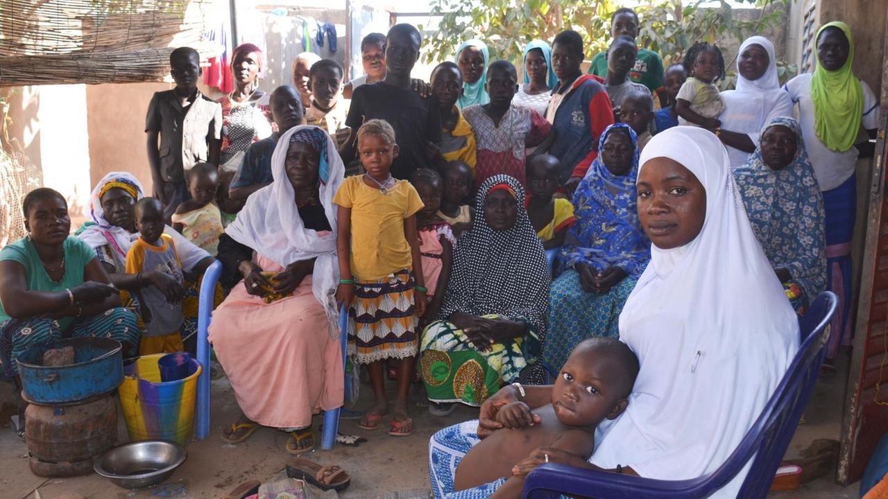 Fatimata Ourba sitzt mit ihren vier Kindern, anderen Familienmitgliedern und Nachbarn auf einem blauen Plastikstuhl und guckt in die Kamera. Sie ist geflüchtet aus der Sahel-Region, lebt jetzt aber in in Pazani - ein Viertel im Norden von Ouagaodougou.
