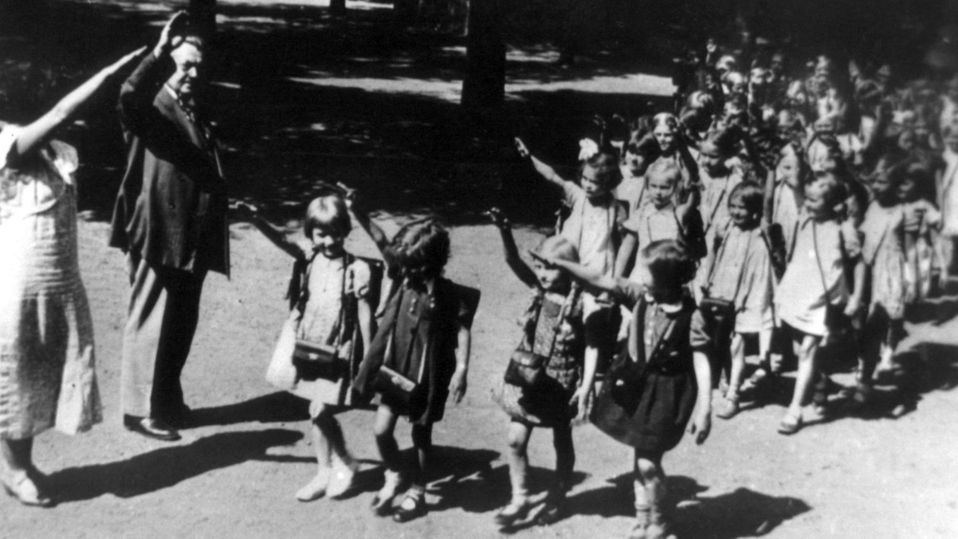 Die rechte Hand zum Hitlergruß erhoben gehen Erstklässler 1933 in Viererreihen an zwei ebenfalls grüßenden Erwachsenen (vermutlich Lehrpersonal) vorbei.